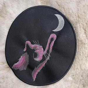May include: A black circular patch with a pink embroidered cat looking up at a silver crescent moon.