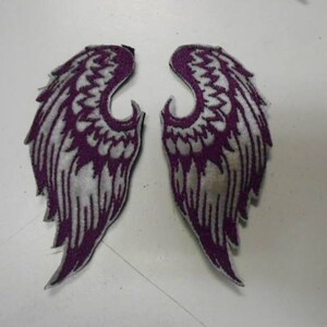 May include: A pair of purple and white embroidered angel wings. The wings are sewn onto a white fabric background.
