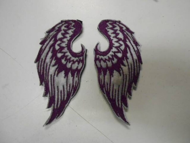 May include: A pair of purple and white embroidered angel wings. The wings are sewn onto a white fabric background.