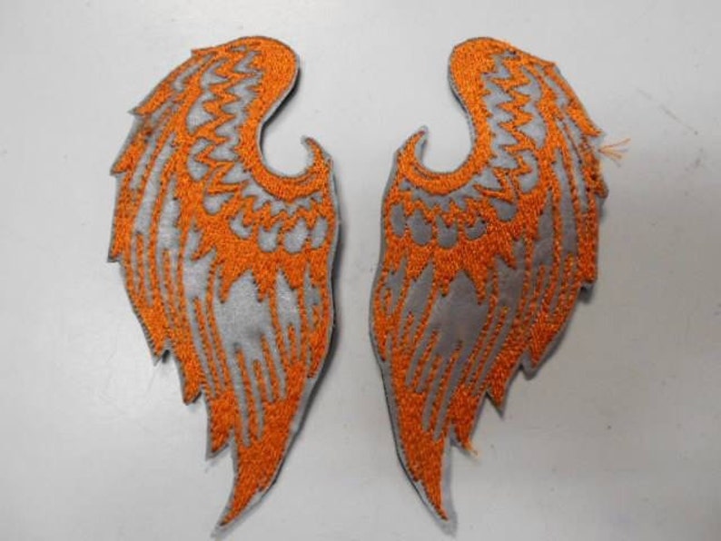 May include: A pair of iron-on patches featuring a silver and orange embroidered design of angel wings.