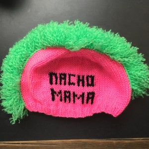 May include: A novelty knit hat with a bright pink base and green faux hair. The words "NACHO MAMA" are embroidered in black on the front of the hat. The hat is designed to resemble a mohawk.