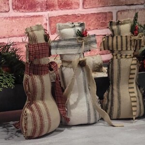 May include: A collection of handmade snowman figurines with fabric bodies and decorative scarves. The snowmen are in various patterns of red, white, and blue, with some adorned with berries and greenery. A festive holiday decoration.