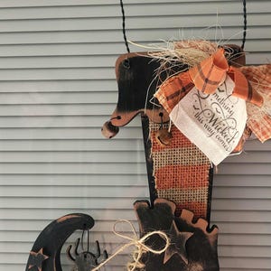 May include: A hanging wooden decoration shaped like a witch boot, painted black with distressed orange edges. It features a burlap stripe, a plaid orange bow, a small spider cutout, wooden stars, and a tag reading "Something Wicked this way comes."