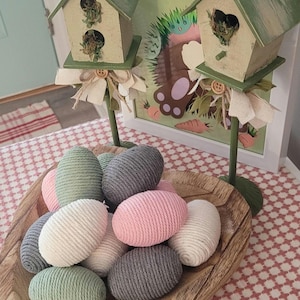 May include: A wooden bowl filled with handmade yarn eggs in shades of pink, gray, and green. The eggs are arranged in a decorative pattern. Two miniature birdhouses with green roofs and white bases are in the background.