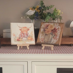May include: Two small wooden canvases on easels. The left canvas features a watercolor painting of a Highland cow wearing a red and white scarf and holding a book. The right canvas features a watercolor painting of a mason jar filled with sunflowers.