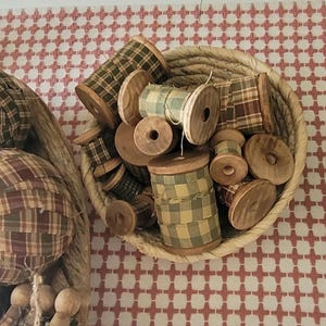 May include: A collection of wooden spools filled with plaid fabric in a woven basket. The spools vary in size and are filled with green, brown, and cream-colored plaid fabric. The basket sits against a red and white patterned background.