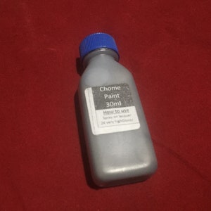 May include: A gray paint bottle with a blue cap, labeled "Chome Paint 30ml." The label includes instructions: "Spray on lacquer" and "2K very highGlossy." The bottle is on a red surface.