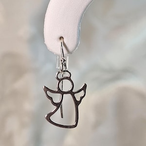 Puede incluir: Un pendiente plateado en forma de &aacute;ngel con cierre de gancho. El pendiente presenta un dise&ntilde;o recortado de un &aacute;ngel con alas y un halo. El metal es brillante y refleja la luz. El pendiente se muestra sobre un fondo blanco.