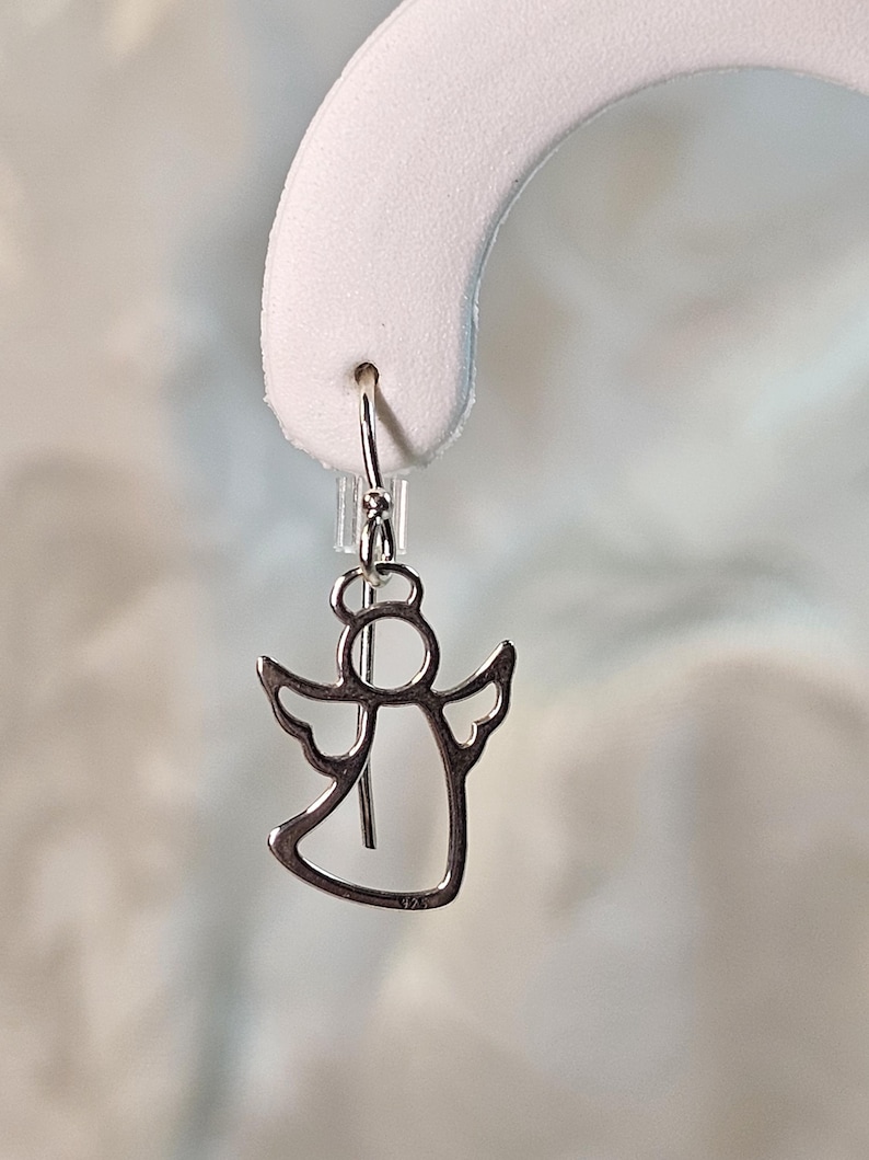 Puede incluir: Un pendiente plateado en forma de &aacute;ngel con cierre de gancho. El pendiente presenta un dise&ntilde;o recortado de un &aacute;ngel con alas y un halo. El metal es brillante y refleja la luz. El pendiente se muestra sobre un fondo blanco.