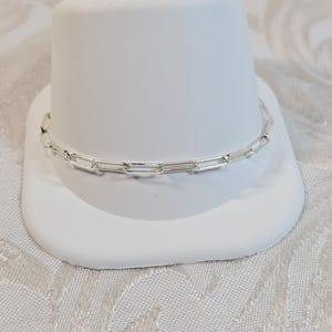May include: A silver paperclip chain necklace displayed on a white pedestal. The necklace is composed of rectangular links. The background is a white and light patterned fabric.