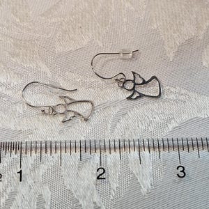 Puede incluir: Un par de pendientes plateados con forma de &aacute;ngel. Los pendientes est&aacute;n hechos de metal fino y tienen cierre de gancho. Los pendientes miden aproximadamente 2,5 cm de largo. Se muestran sobre una superficie blanca texturizada.