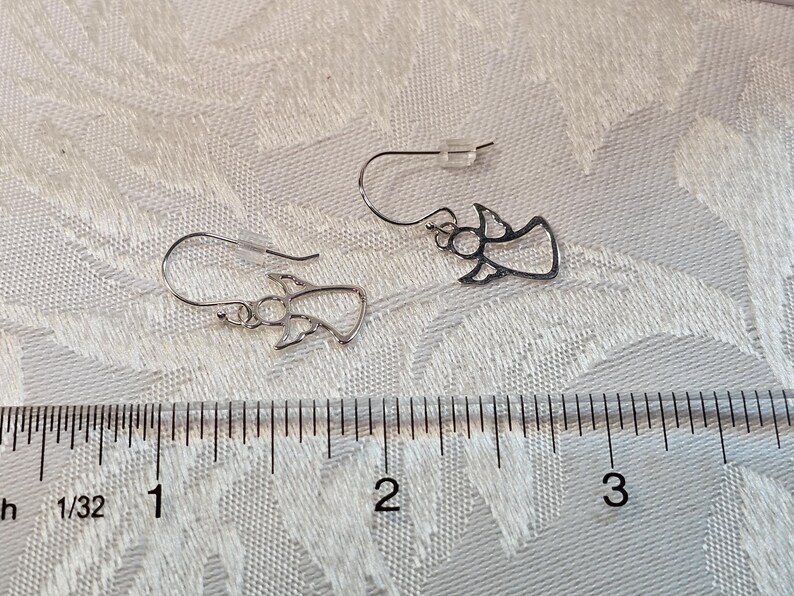 Puede incluir: Un par de pendientes plateados con forma de &aacute;ngel. Los pendientes est&aacute;n hechos de metal fino y tienen cierre de gancho. Los pendientes miden aproximadamente 2,5 cm de largo. Se muestran sobre una superficie blanca texturizada.