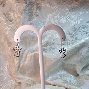 Puede incluir: Un par de pendientes de &aacute;ngel plateados colgando de un expositor blanco. Los pendientes presentan un dise&ntilde;o de silueta de &aacute;ngel simple. El fondo es una tela estampada con un dise&ntilde;o floral.