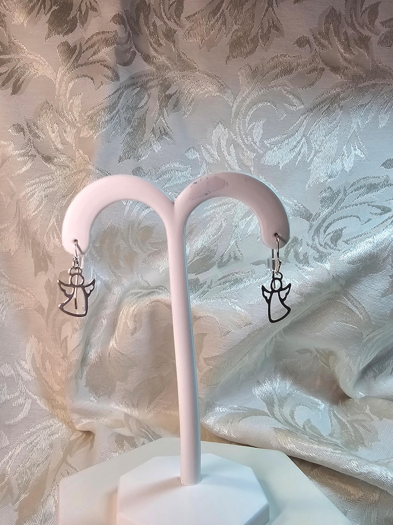 Puede incluir: Un par de pendientes de &aacute;ngel plateados colgando de un expositor blanco. Los pendientes presentan un dise&ntilde;o de silueta de &aacute;ngel simple. El fondo es una tela estampada con un dise&ntilde;o floral.