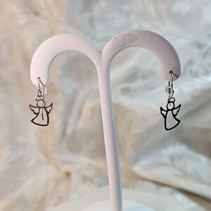 Puede incluir: Un par de pendientes plateados con forma de &aacute;ngel. Cada pendiente presenta un dise&ntilde;o de &aacute;ngel recortado suspendido de un peque&ntilde;o gancho. Los pendientes se exhiben en un soporte blanco para joyas sobre un fondo suave y estampado.
