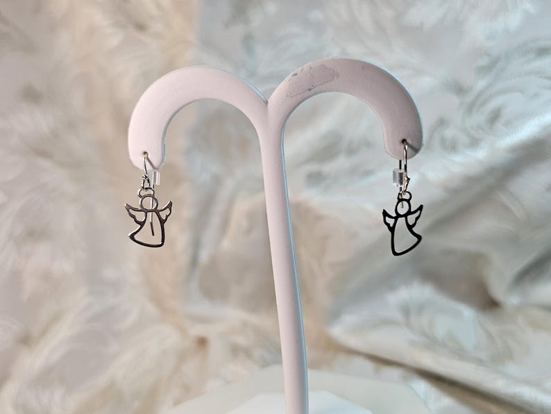 Puede incluir: Un par de pendientes plateados con forma de &aacute;ngel. Cada pendiente presenta un dise&ntilde;o de &aacute;ngel recortado suspendido de un peque&ntilde;o gancho. Los pendientes se exhiben en un soporte blanco para joyas sobre un fondo suave y estampado.