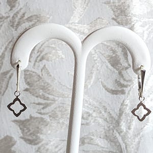 May include: A pair of silver earrings with a delicate, openwork design in the shape of a four-leaf clover. The earrings are hanging from a simple hoop.