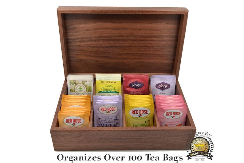 May include: A wooden tea bag organizer with a hinged lid. The organizer is filled with various tea bags, including Bigelow Cozy Chamomile, Yogi, and Red Rose. The text "Organizes Over 100 Tea Bags" is printed on the front of the organizer.