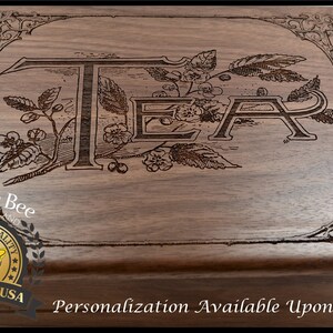 May include: A wooden tea box with an engraved design of the word "TEA" surrounded by floral motifs. The box has a dark brown wood finish and a gold label with the words "Designer Bee" and "Premium Quality" on it. The label also has a bee illustration and the words "Made in USA". The text "Personalization Available Upon Checkout" is printed on the bottom of the box.