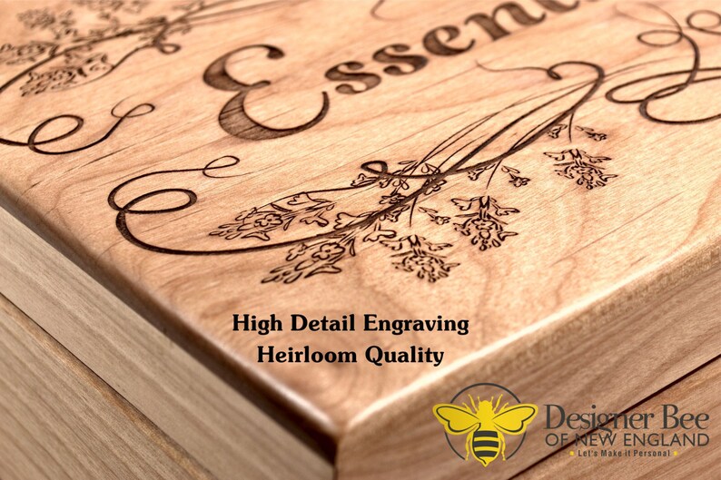 May include: A wooden box with a detailed engraving of the word "Essence" and floral designs. The box is made of light wood and has a natural finish. The text "High Detail Engraving Heirloom Quality" is printed on the box. A bee logo with the text "Designer Bee of New England Let's Make it Personal" is printed on the bottom right corner of the box.