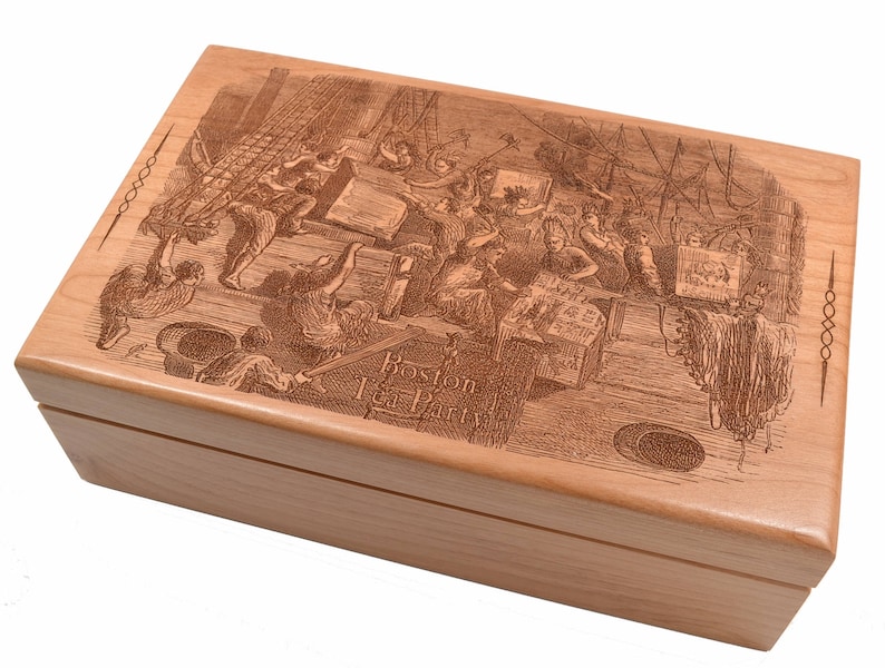 Op de afbeelding: Een houten doos met een gegraveerd beeld van de Boston Tea Party. De doos is lichtbruin en heeft een scharnierend deksel. De gravure toont een groep mensen die theekisten in het water gooien.