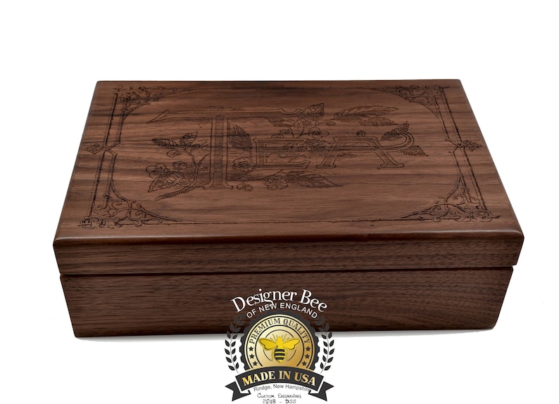 May include: A wooden box with an engraved design of a bee and flowers. The box is closed and has a label on the front that reads "Designer Bee of New England" and a logo of a bee with the words "Premium Quality Made in USA".