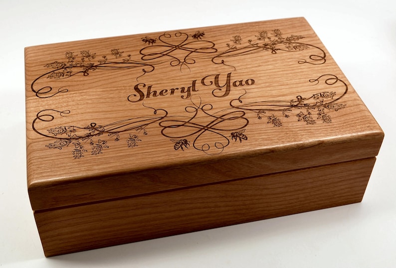 May include: A wooden box with an engraved design of flowers and bees. The box is personalized with the name "Sheryl Yao".
