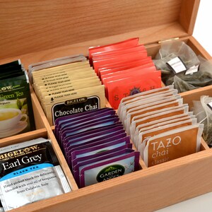 Op de afbeelding: Een houten theedoos met meerdere vakken gevuld met verschillende theezakjes. De theezakjes zijn gesorteerd op type, waaronder groene thee, chocolade chai, Earl Grey en andere. De doos is gemaakt van licht hout en heeft een scharnierend deksel.