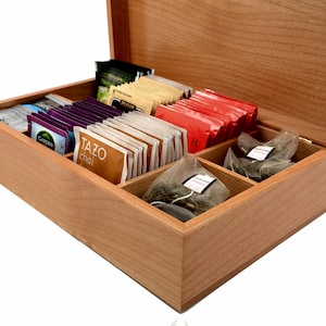 Op de afbeelding: Een houten theedoos met een scharnierend deksel, open om zes vakken te onthullen die gevuld zijn met verschillende theezakjes. De vakken zijn gelabeld met de theemerken "Tazo chai", "Garden" en "H", en de theezakjes zijn in verschillende kleuren en ontwerpen.