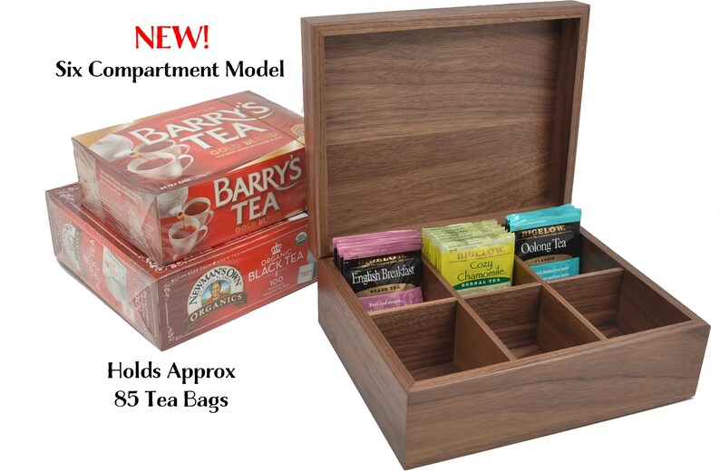 May include: A wooden tea box with six compartments, open to reveal tea bags. The box is made of dark wood and has a lid. The tea bags are in individual compartments and are labeled with different tea flavors. The box is labeled "NEW! Six Compartment Model" and "Holds Approx 85 Tea Bags".