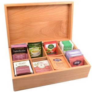 Op de afbeelding: Een houten theedoos met een deksel, open om negen vakken te onthullen gevuld met verschillende theezakjes. De theezakjes zijn gelabeld met merknamen zoals Bigelow, Garden Andes, Salada en Yogi.