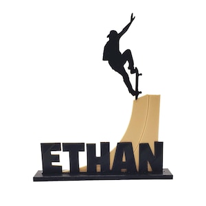 May include: A black silhouette of a skateboarder mid-air, performing a trick on a ramp. The ramp is tan, and the name "ETHAN" is displayed in black block letters at the base. The entire piece is mounted on a black base.
