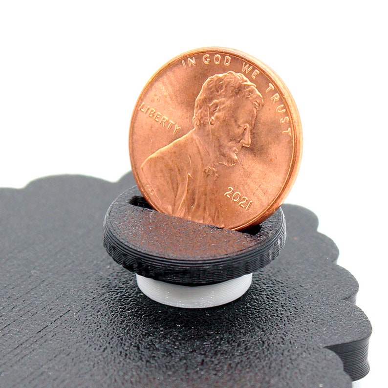 May include: A black plastic coin holder with a white base. A copper-colored 2021 US penny is resting in the holder. The penny has the words "IN GOD WE TRUST" and "LIBERTY" on it.