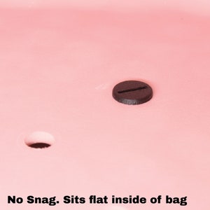May include: A black plastic snap button with a single slot. The button is sitting on a pink surface. "No Snag. Sits flat inside of bag" is written below the button.