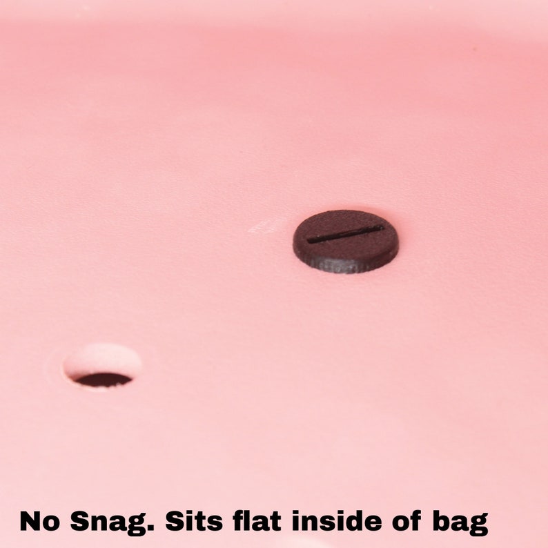 May include: A black plastic snap button with a single slot. The button is sitting on a pink surface. "No Snag. Sits flat inside of bag" is written below the button.