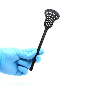 Könnte beinhalten: Ein schwarzer Lacrosse-Schläger mit einem schwarzen Netz. Der Schläger wird in einer behandschuhten Hand gehalten.
