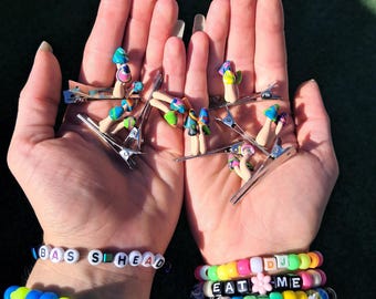 May include: A collection of silver metal hair clips adorned with colorful mushroom-shaped charms. The image also shows multiple beaded bracelets spelling out "BAS S L HEAD", "DJ", "EAT ME", and "LUC". The bracelets are in various colors.