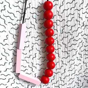 May include: A necklace featuring a combination of red spherical beads and light pink geometric shapes. The necklace hangs on a black cord against a white fabric background with a black wavy pattern.