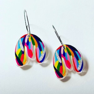 May include: A pair of colorful abstract earrings. Each earring features a clear, curved shape with a vibrant pattern of rainbow-colored brushstrokes and white dotted accents. The earrings hang from silver-colored hoops.
