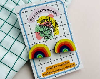 Op de afbeelding: Een paar regenboogkleurige houten oorbellen. De oorbellen zijn halfrond en hebben een geel, oranje, rood, blauw, groen en paars ontwerp. De oorbellen liggen op een witte kaart met de tekst "rainbowrosemakes" en een social media-handle.