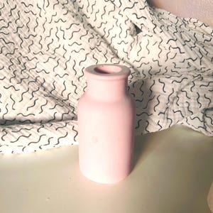 May include: A light pink ceramic vase with a rounded shape. The vase is sitting on a white surface with a black and white patterned fabric behind it.