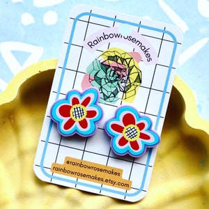 May include: A pair of colourful flower-shaped earrings with a red centre, blue and purple petals, and a black and white chequered pattern. The earrings are displayed on a white card with the text "Rainbowrosemakes" and "@rainbowrosemakes rainbowrosemakes.etsy.com".