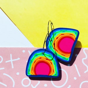 May include: A pair of rainbow hoop earrings with a blue outline. The earrings are made of polymer clay and have a layered rainbow design with a pink center.