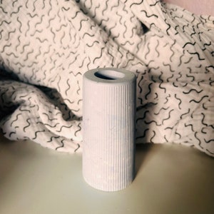 May include: A light blue, ribbed ceramic vase with a round opening at the top. The vase is sitting on a white surface.