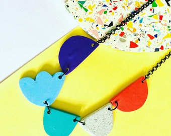 May include: A colorful necklace featuring geometric shapes in blue, red, turquoise, and speckled white, connected by a black chain. The necklace is displayed on a yellow and white background with a terrazzo-style accent.