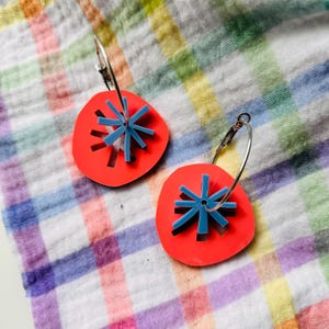 May include: A pair of red hoop earrings with a blue starburst design. The earrings are on a checkered fabric background.