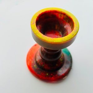May include: A small, colorful resin cup with a yellow rim and a red, green, and black speckled base. The cup is designed to hold a small object, such as a ring or a key.