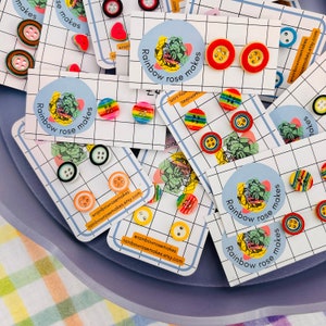 May include: A collection of colorful button earrings on display. The earrings feature various designs, including solid colors, stripes, and patterns. Each pair is mounted on a small card with the text "Rainbow rose makes".