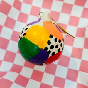 May include: A white ceramic bauble with a geometric pattern in shades of yellow, green, purple, red, orange, and black. The bauble has a gold string for hanging.