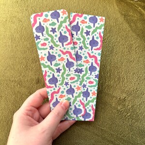 Può includere: Due segnalibri colorati con un motivo a tema oceanico. Il design presenta pesci luna viola, pesci arancioni, alghe verdi, linee ondulate rosa e stelle marine blu su sfondo bianco. I segnalibri sono tenuti in una mano.